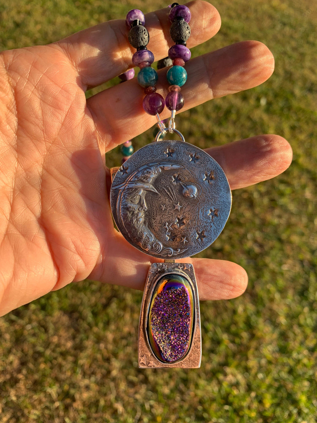 Hand holding a silver medallion with star design and a purple gemstone pendant against a grassy background.