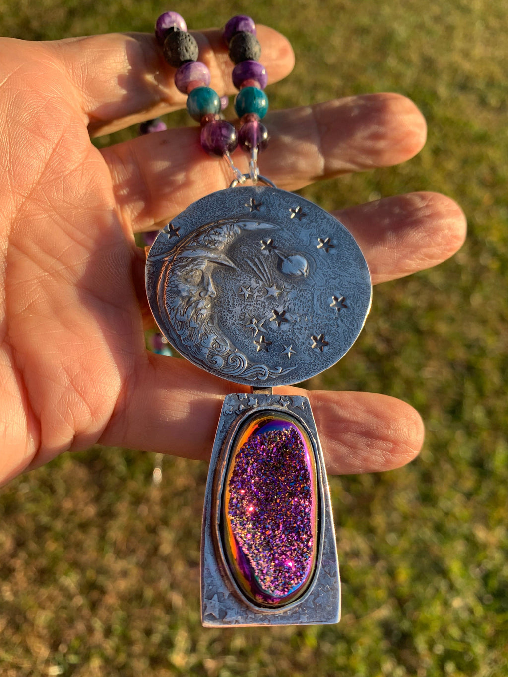 Hand holding a silver pendant with star and moon design, a purple druzy stone, and a beaded chain against a grassy background.