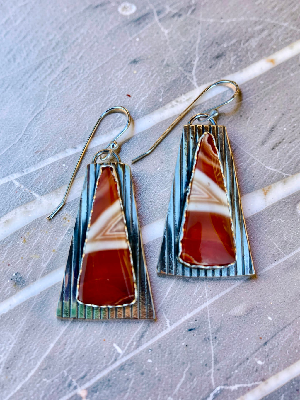 One-of-a-kind Echoes in the Stone earrings by Christine Keller, featuring red and cream banded agate gemstones set on elongated textured silver backplates with silver ear wires.