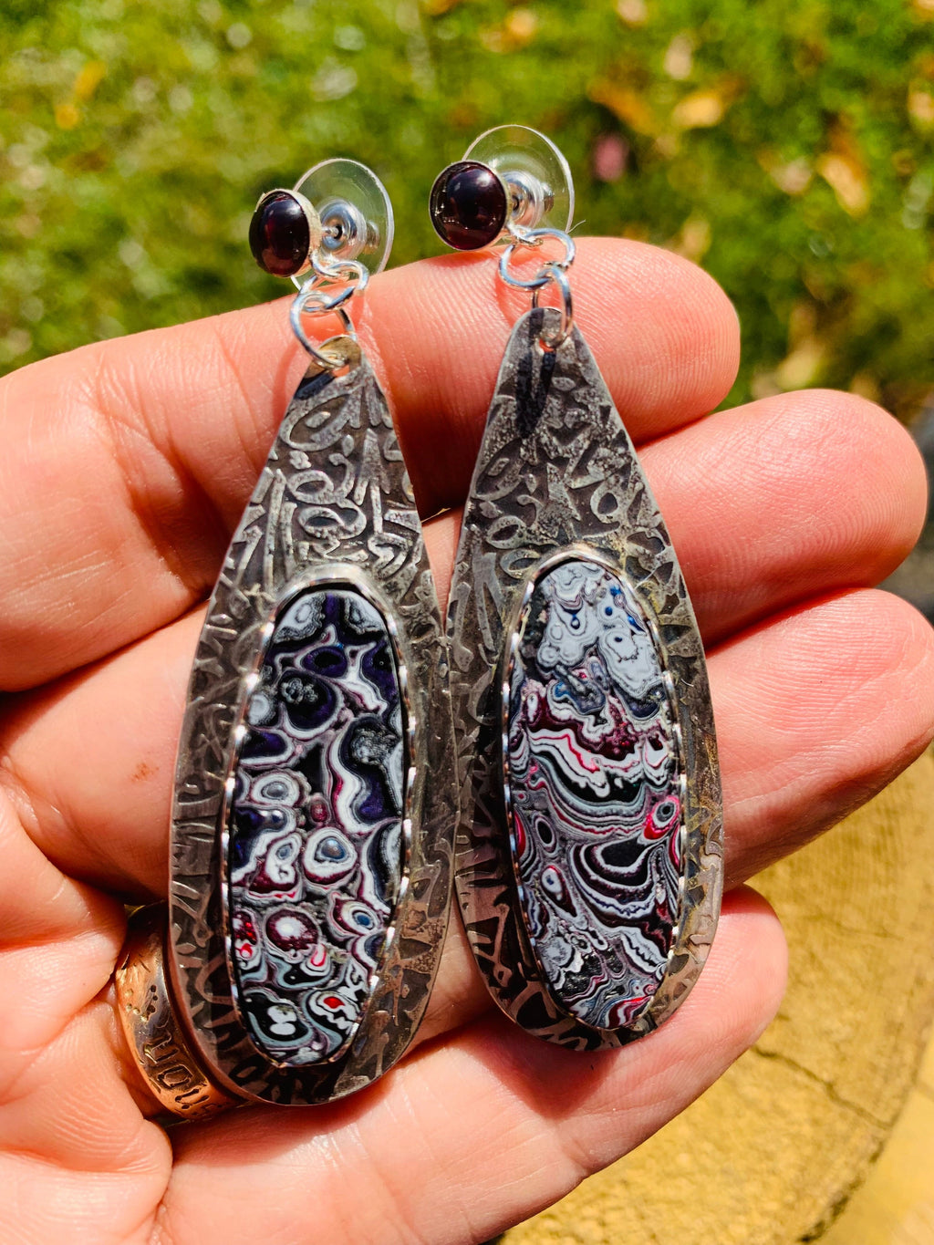 Pair of silver earrings with intricate designs held in a hand against a blurred natural background.