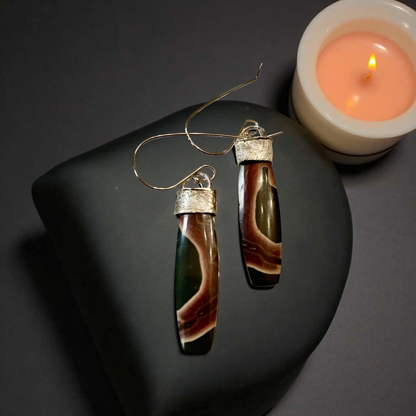 Pair of jasper earrings on a dark stone surface with a candle in the background.
