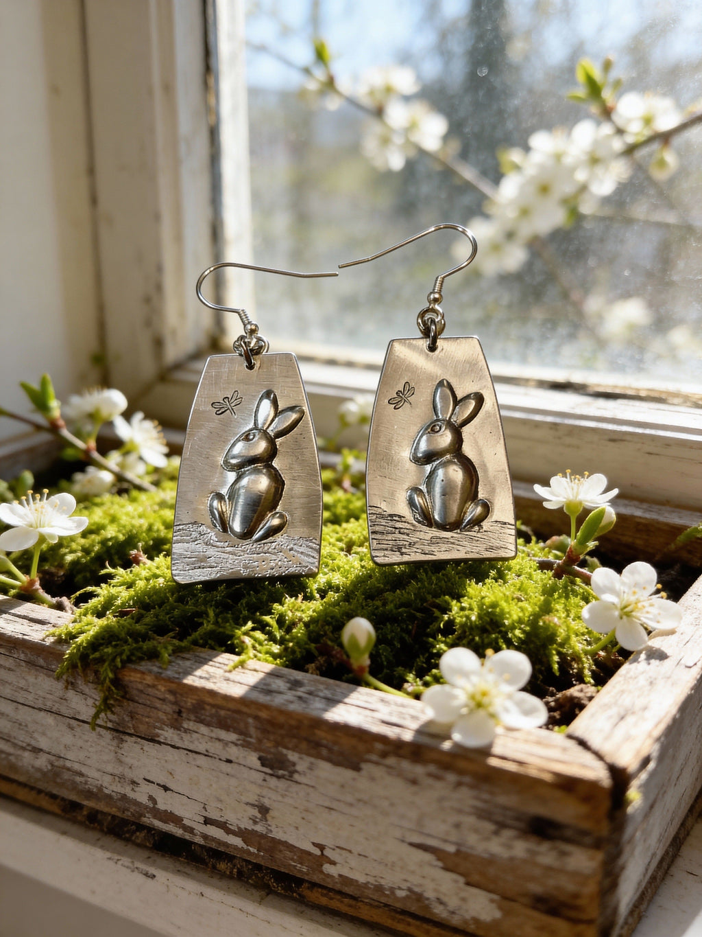 Pair of silver rabbit-shaped earrings on a wooden surface with flowers and moss.