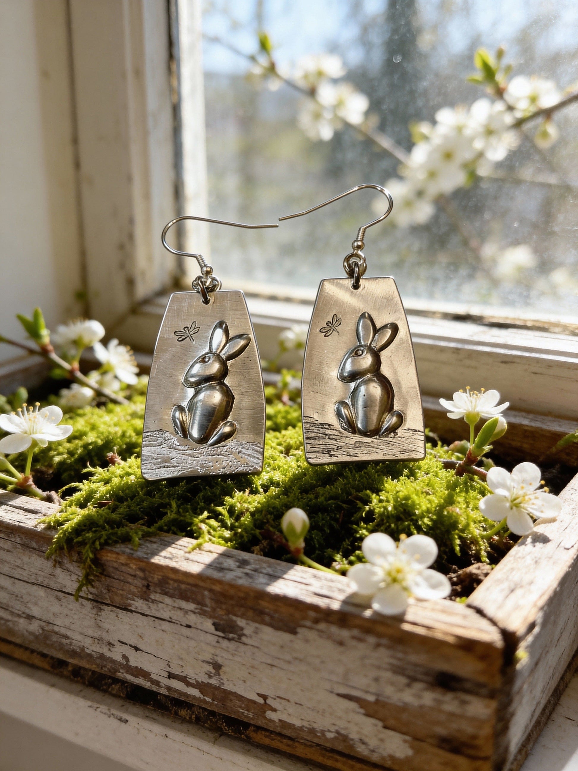 Pair of silver rabbit-shaped earrings on a wooden surface with flowers and moss.