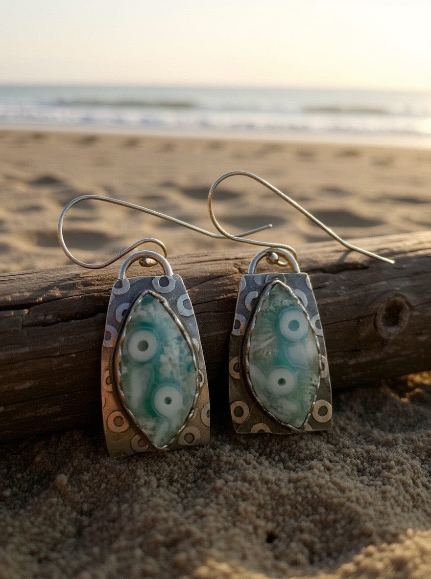 Pair of earrings with  blue green and silver design on a wooden log by the beach.