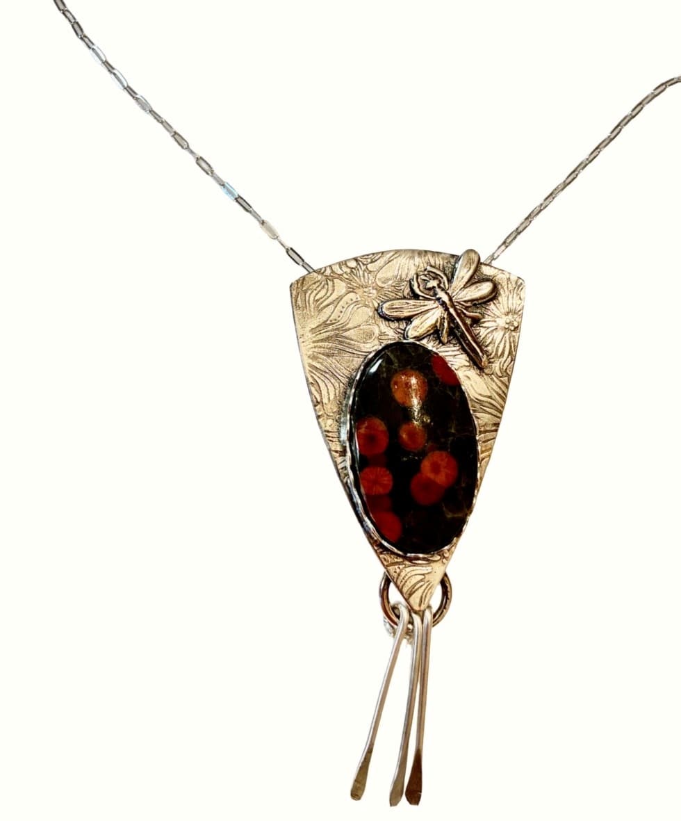 A silver pendant necklace featuring a shield-shaped textured metal frame, a small dragonfly charm, and an oval dark red and black stone centerpiece, shown on a plain white background.