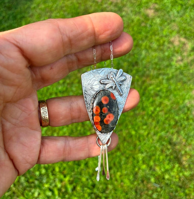A hand holding a silver pendant necklace outdoors with a green grass background. The pendant features a dragonfly motif, a silver shield background, and an oval black and orange stone inlay.