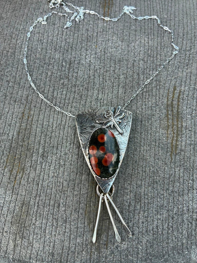 A silver pendant necklace featuring a shield-shaped textured silver frame, a small dragonfly motif, and a central oval black stone with red spots, resting on a gray textured surface.