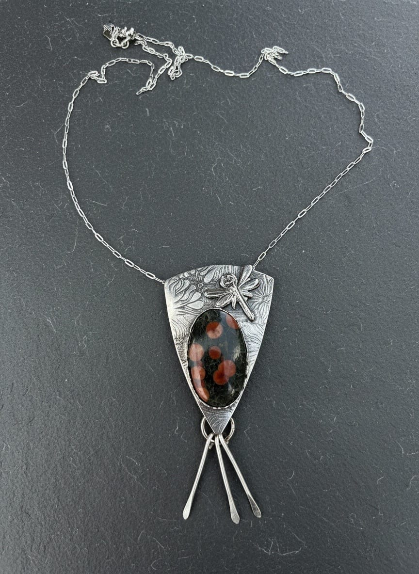 A silver pendant necklace featuring a shield-shaped silver frame with a central oval cabochon stone in black and orange-brown, plus three dangling silver bars below, displayed on a dark gray textured surface.