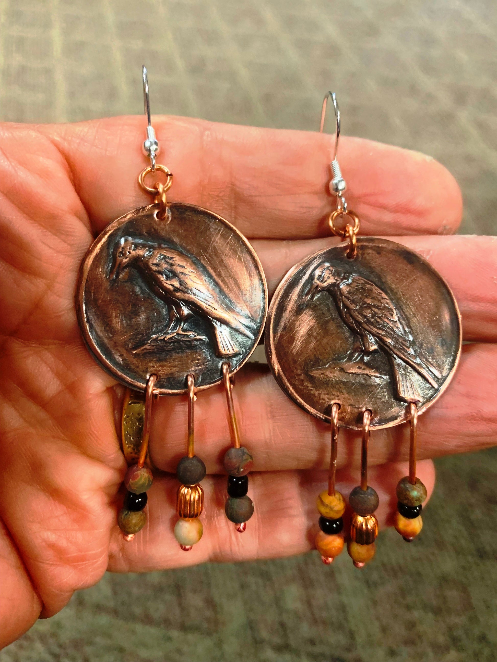 Hand holding a pair of coin-shaped earrings with bird designs and bead accents.