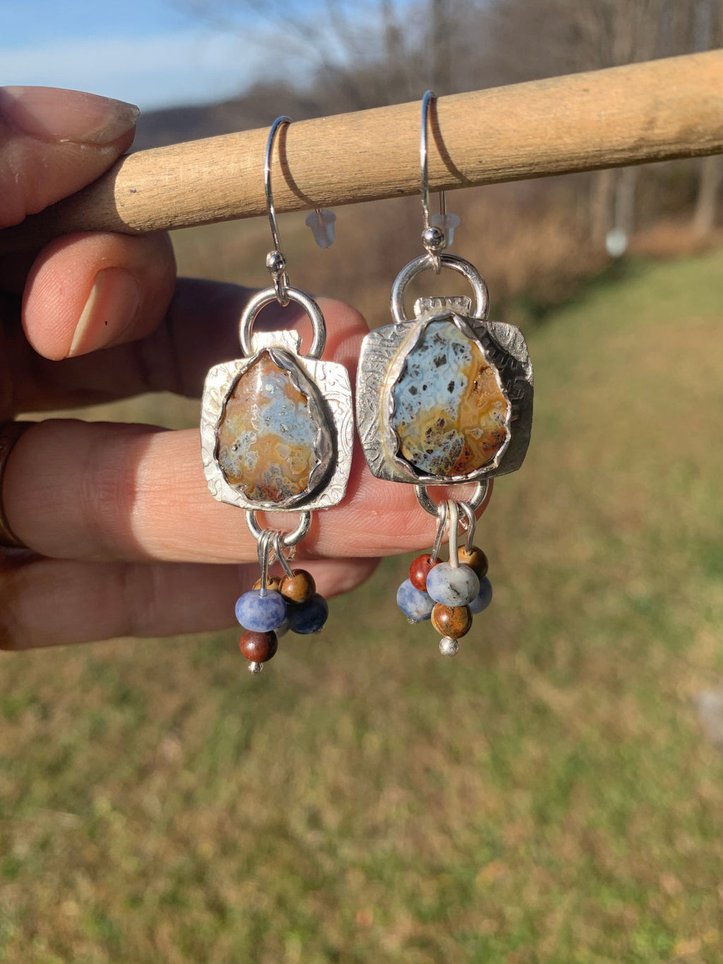   Silver earrings with stone and bead designs held by a hand against a blurred natural background.
