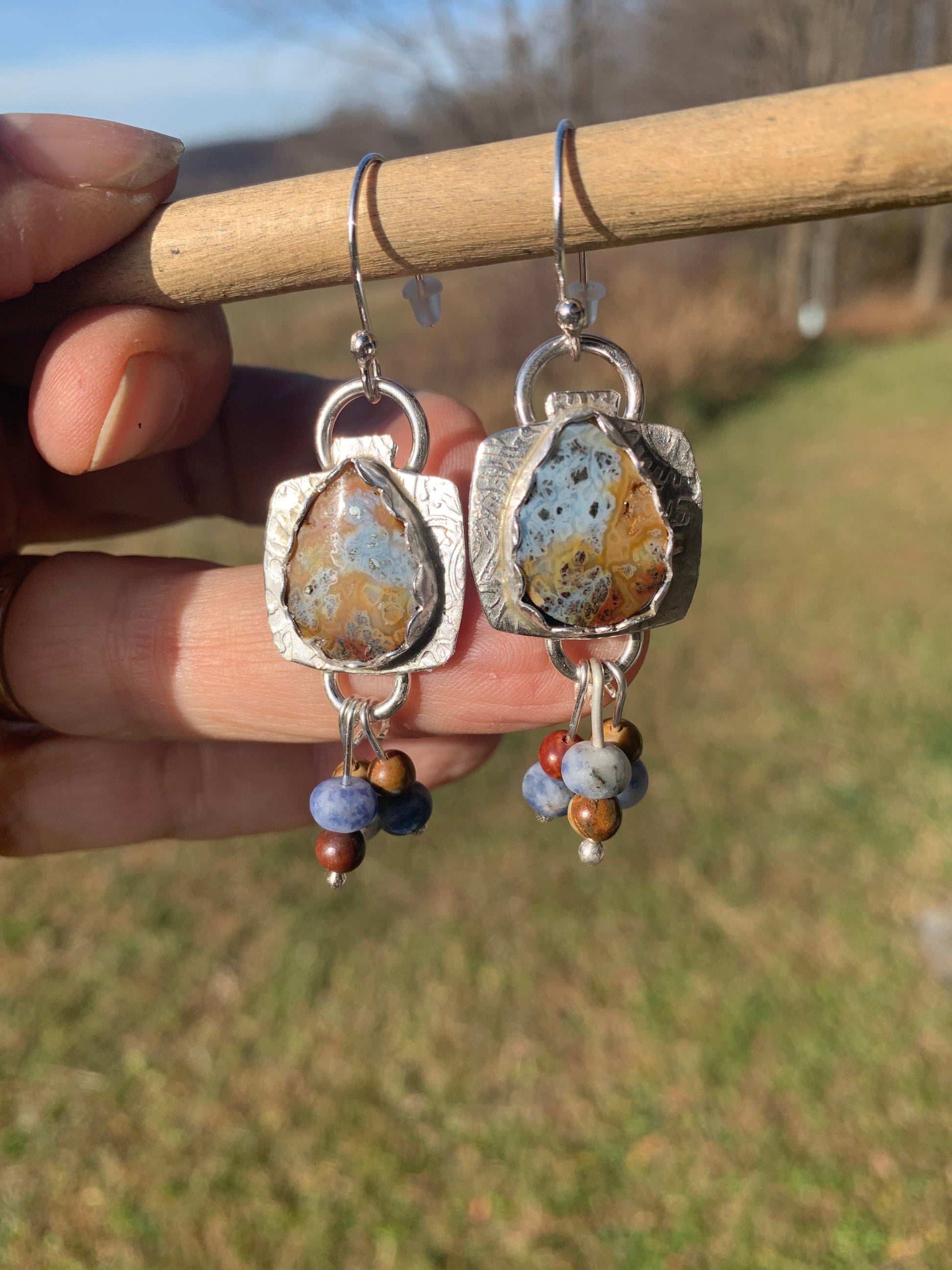   Silver earrings with stone and bead designs held by a hand against a blurred natural background.