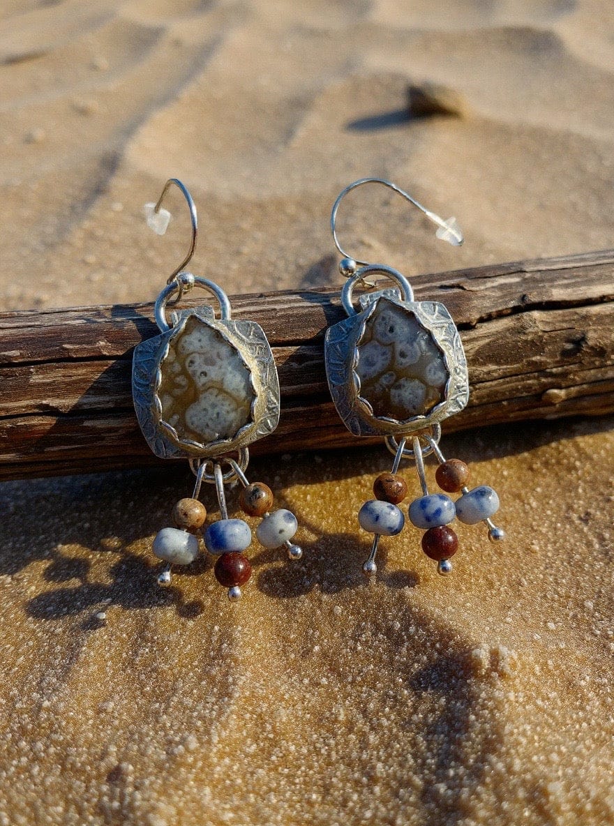 Pair of silver earrings with natural stone and bead charms on a sandy surface.