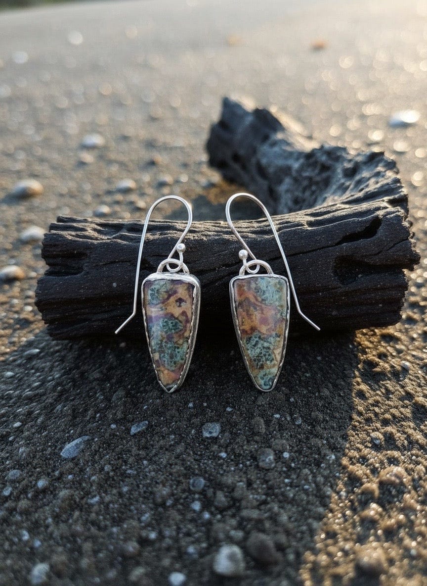 Silver earrings with turquoise stones on a piece of driftwood on a beach.