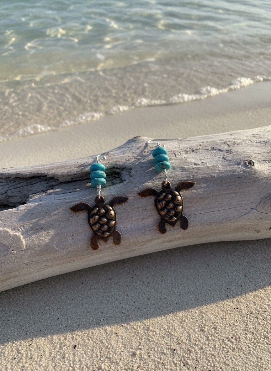 Tortoise-shaped earrings with turquoise beads on a piece of driftwood by the beach.