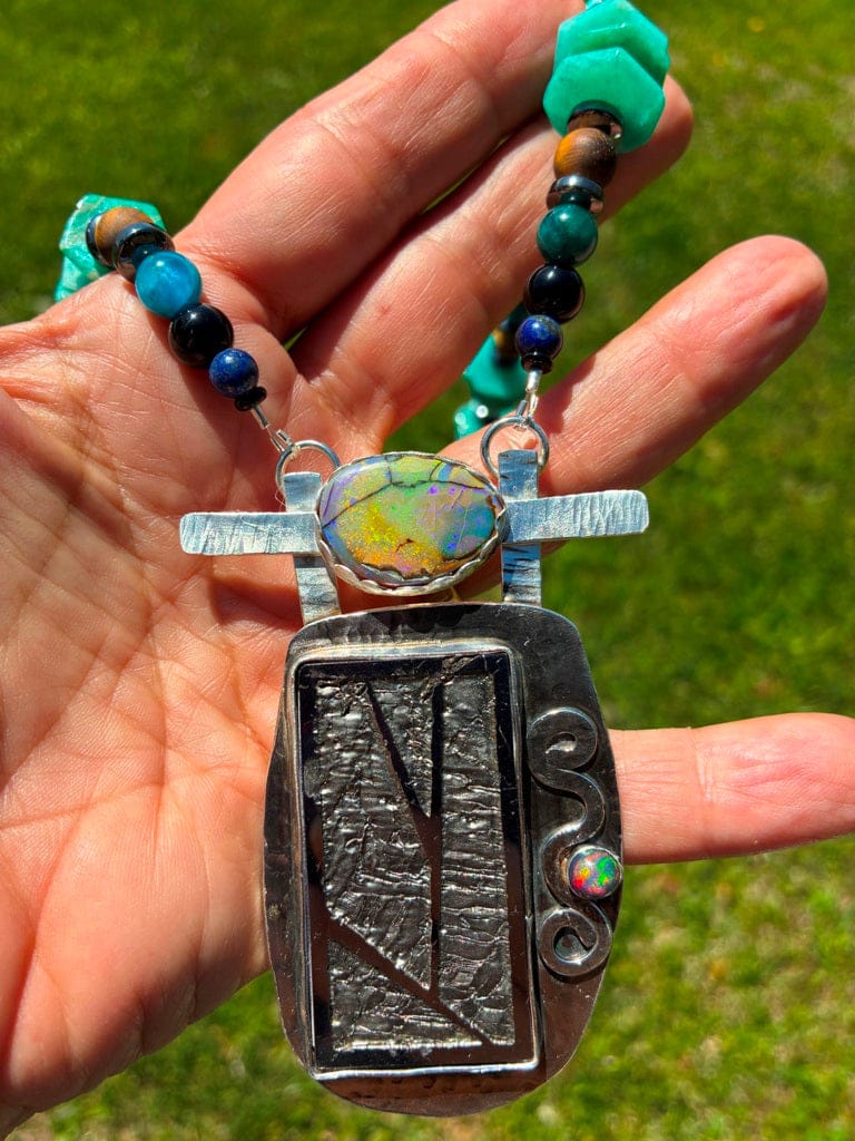 Hand holding a necklace with a silver pendant and colorful opals against a grassy background