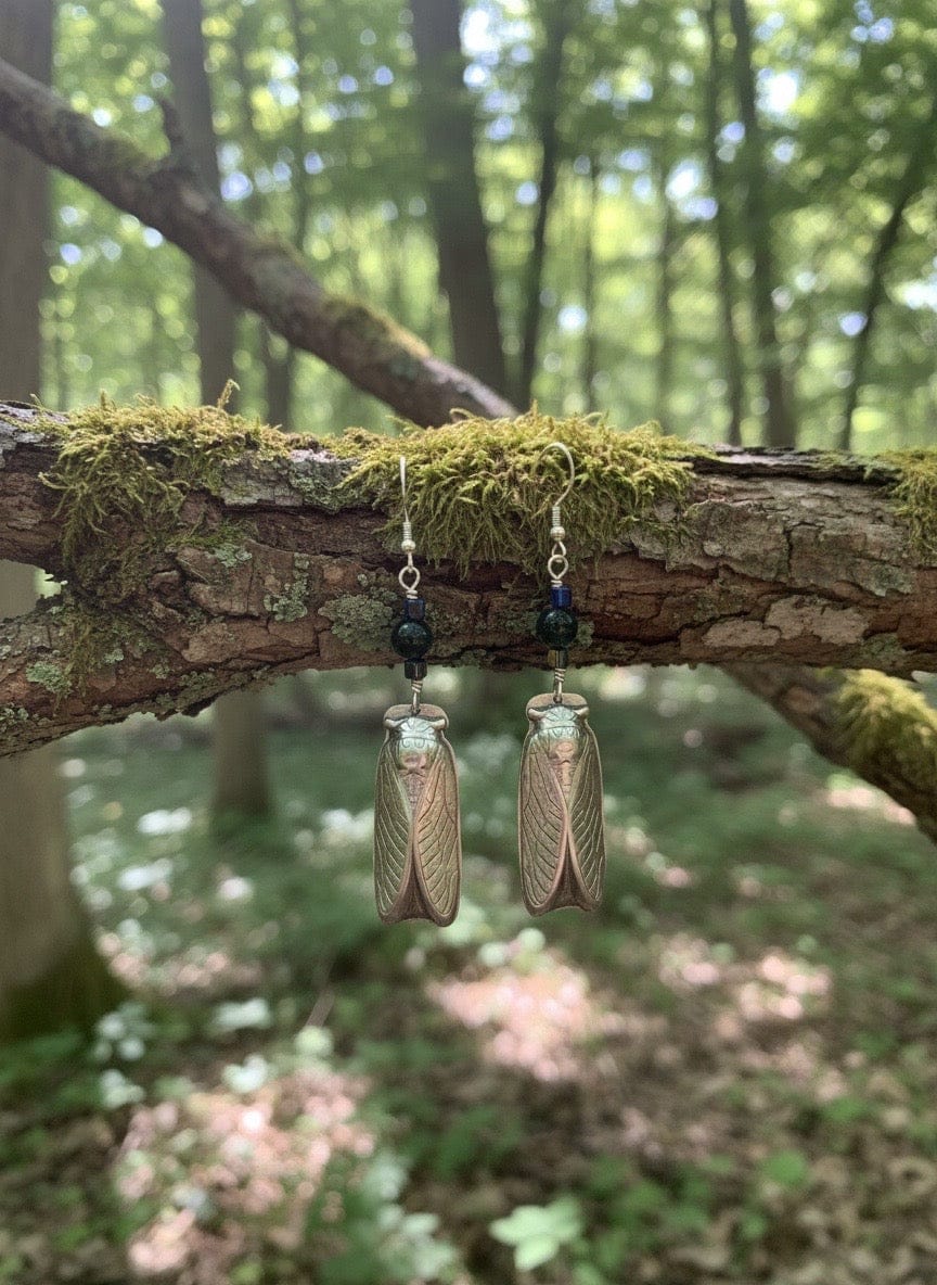 Pair of cicada-themed earrings hanging on a branch with moss in a forest setting