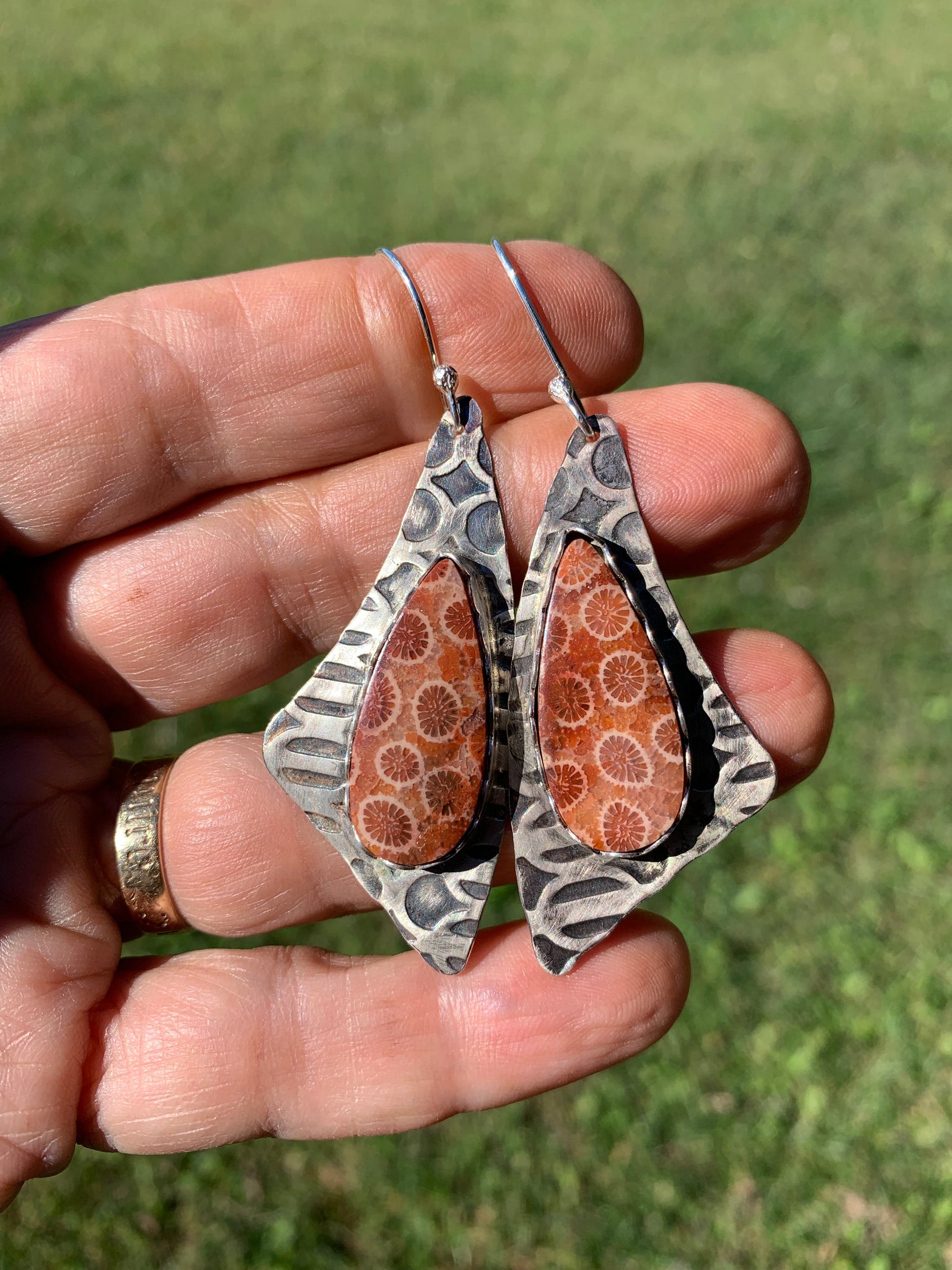 Hand holding a pair of earrings with a patterned design against a grassy background